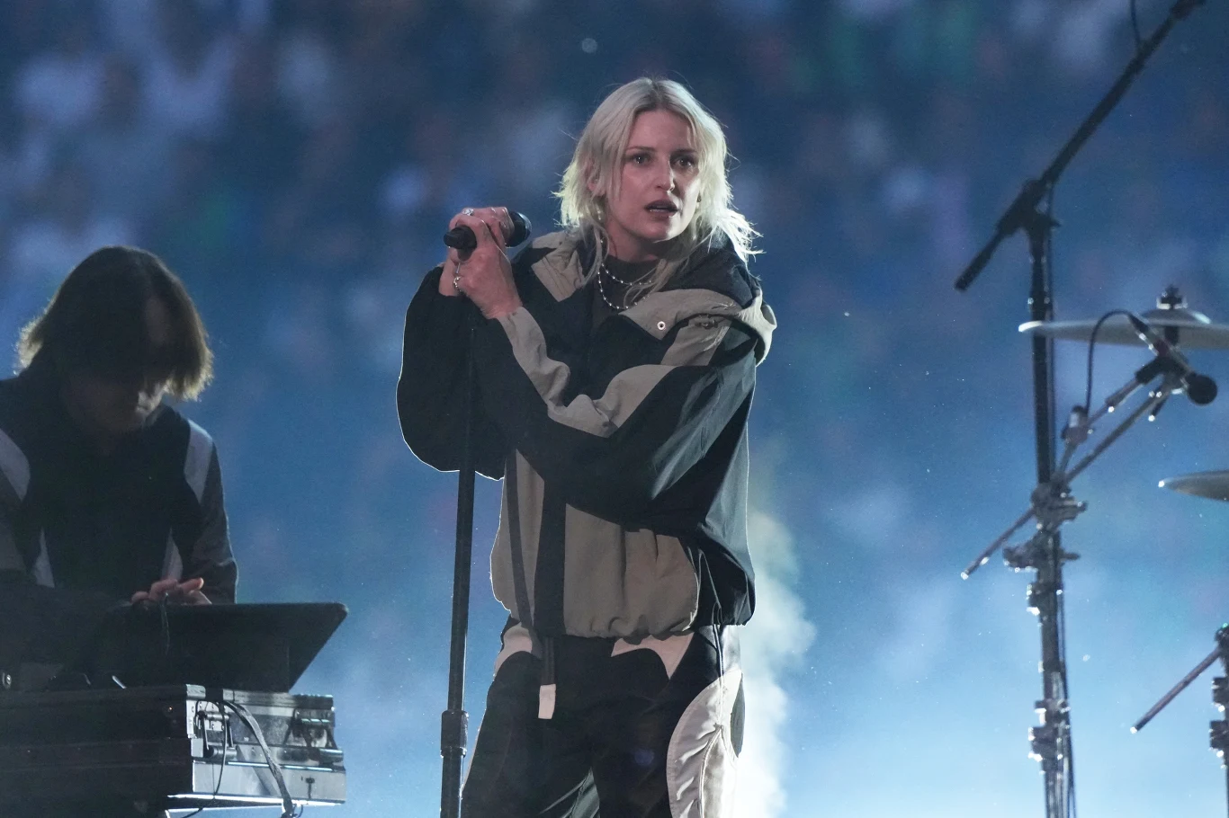 Linkin Park Piosenkarka z jasnymi włosami trzyma mikrofon i występuje na scenie przy świetle reflektorów, obok niej znajduje się osoba obsługująca sprzęt muzyczny, w tle widownia i rozmyte światła.