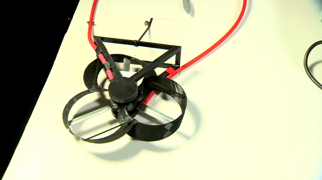 Napęd do dronów stworzony przez Polaków ma nietypowe śmigło Nowoczesny model drona z trzema otwartymi wirnikami zamocowany na biurku, wykonany z lekkich materiałów, widoczny czerwony przewód zasilający urządzenie.