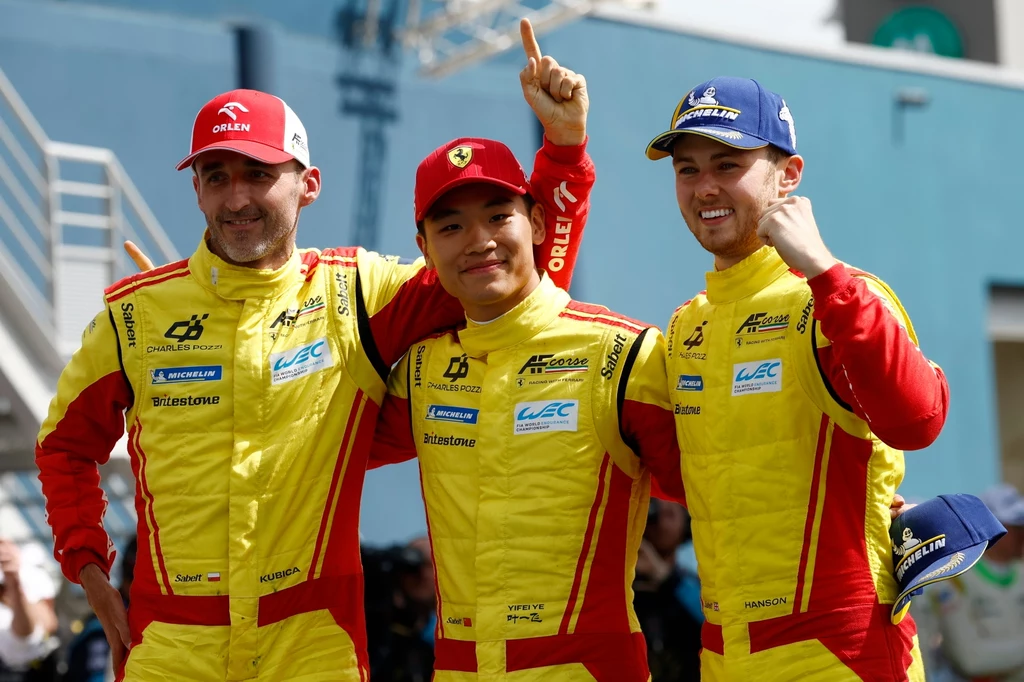 Robert Kubica i jego koledzy z zespołu na mecie Le Mans Trzech kierowców wyścigowych w żółto-czerwonych kombinezonach drużynowych pozuje do wspólnego zdjęcia, jeden z nich unosi palec wskazujący w geście zwycięstwa, wszyscy się uśmiechają, w tle widoczne elementy toru wyścigowego.