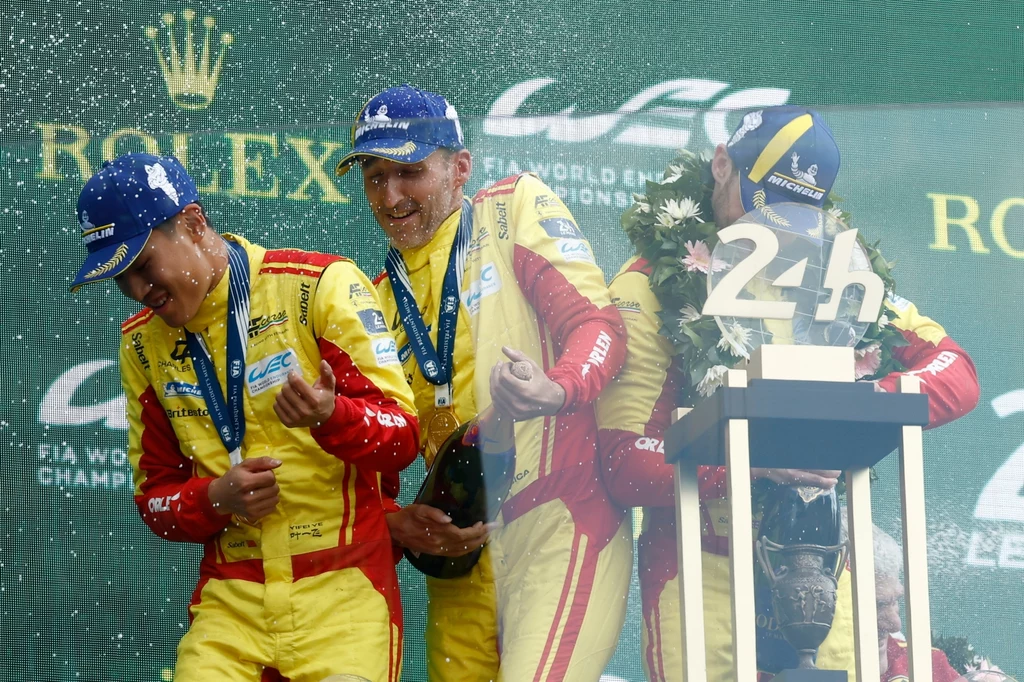 Zwycięski zespół na mecie Le Mans 24h Trzech kierowców wyścigowych w żółto-czerwonych kombinezonach świętuje zwycięstwo na podium, oblewając się szampanem, na szyjach medale, obok puchar oraz wieniec laurowy.