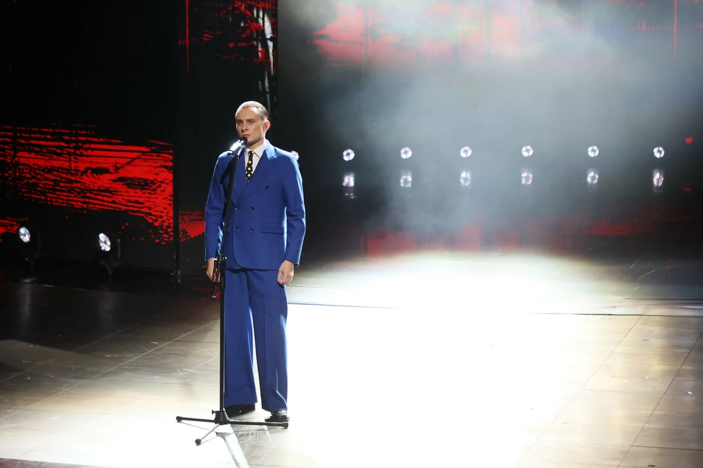 Mężczyzna w niebieskim garniturze stoi samotnie na scenie przy mikrofonie, w tle rozświetlone światła i czerwona iluminacja, otoczony lekką mgłą scenicznego dymu.