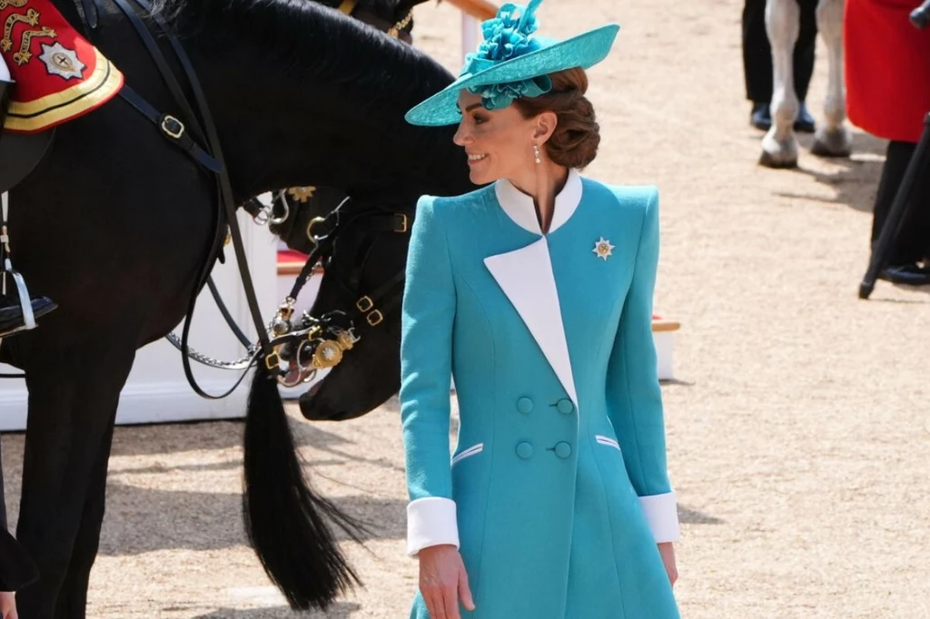Strój księżnej Walii był ukłonem w stronę teściowej Elegancka kobieta w turkusowym płaszczu i kapeluszu stoi na zewnątrz przy czarnym koniu, w tle osoby w strojach ceremonialnych; kobieta uśmiecha się, a koń pochyla głowę w jej stronę.