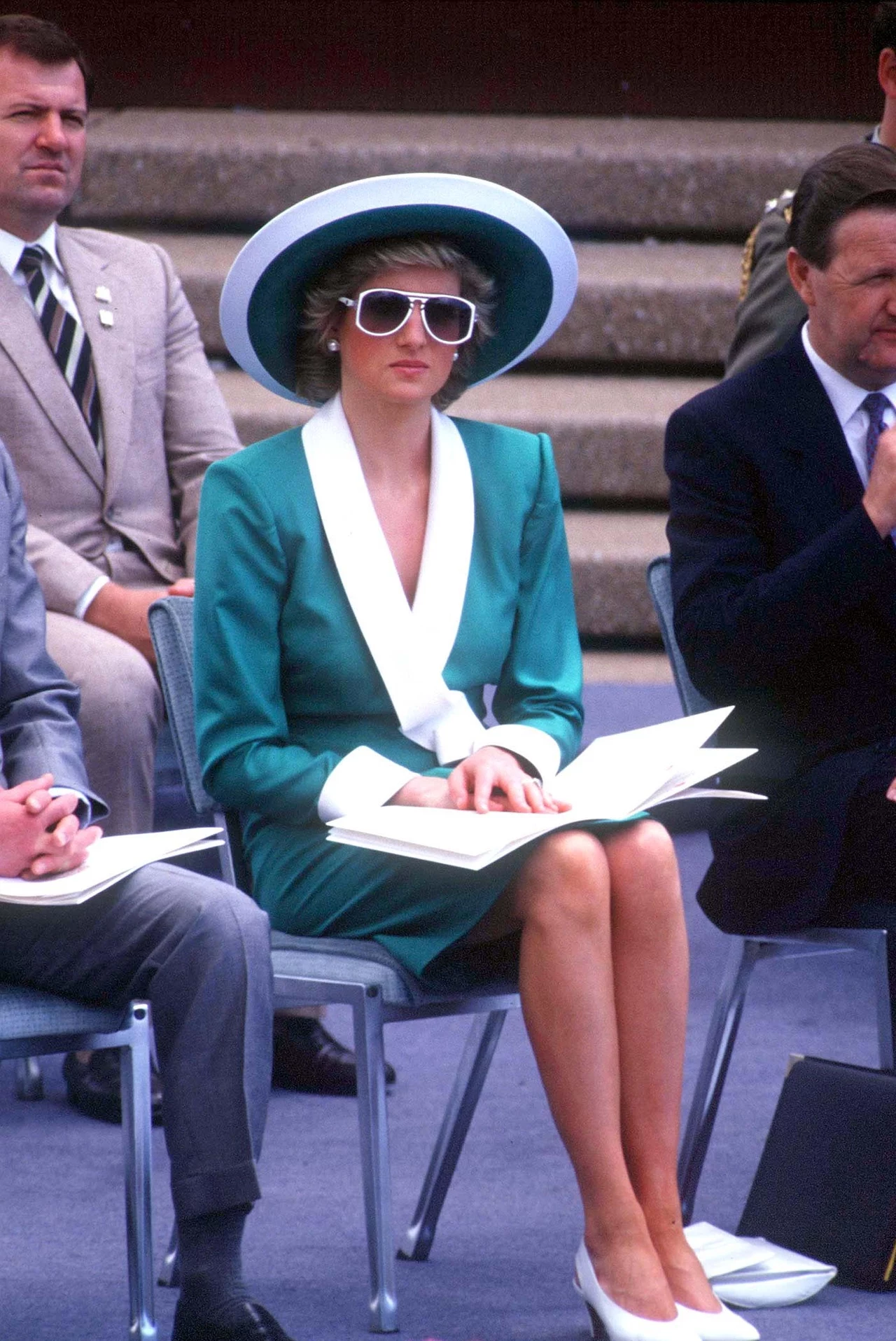księżna Diana i jej styl wciąż są inspiracją dla wielu osób, w tym dla księżnej Kate Kobieta w eleganckim turkusowym stroju z białymi akcentami siedzi na krześle, trzymając na kolanach dokumenty; ma na głowie ozdobny kapelusz i duże okulary przeciwsłoneczne, obok niej znajdują się inni uczestnicy formalnego wydarzenia.