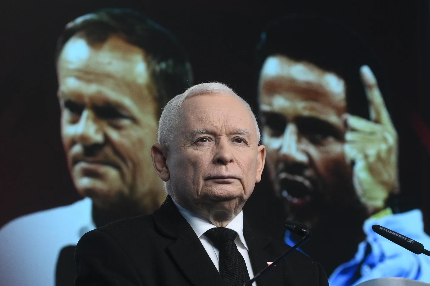Taśmy są pierwszym krokiem do powrotu do władzy Portret starszego mężczyzny w garniturze na pierwszym planie, w tle widoczne wyraźne sylwetki dwóch innych mężczyzn, z których jeden gestykuluje podniesioną ręką i wyrazem twarzy sugerującym emocje, całość sprawia wrażenie momentu politycznego wystąpie...