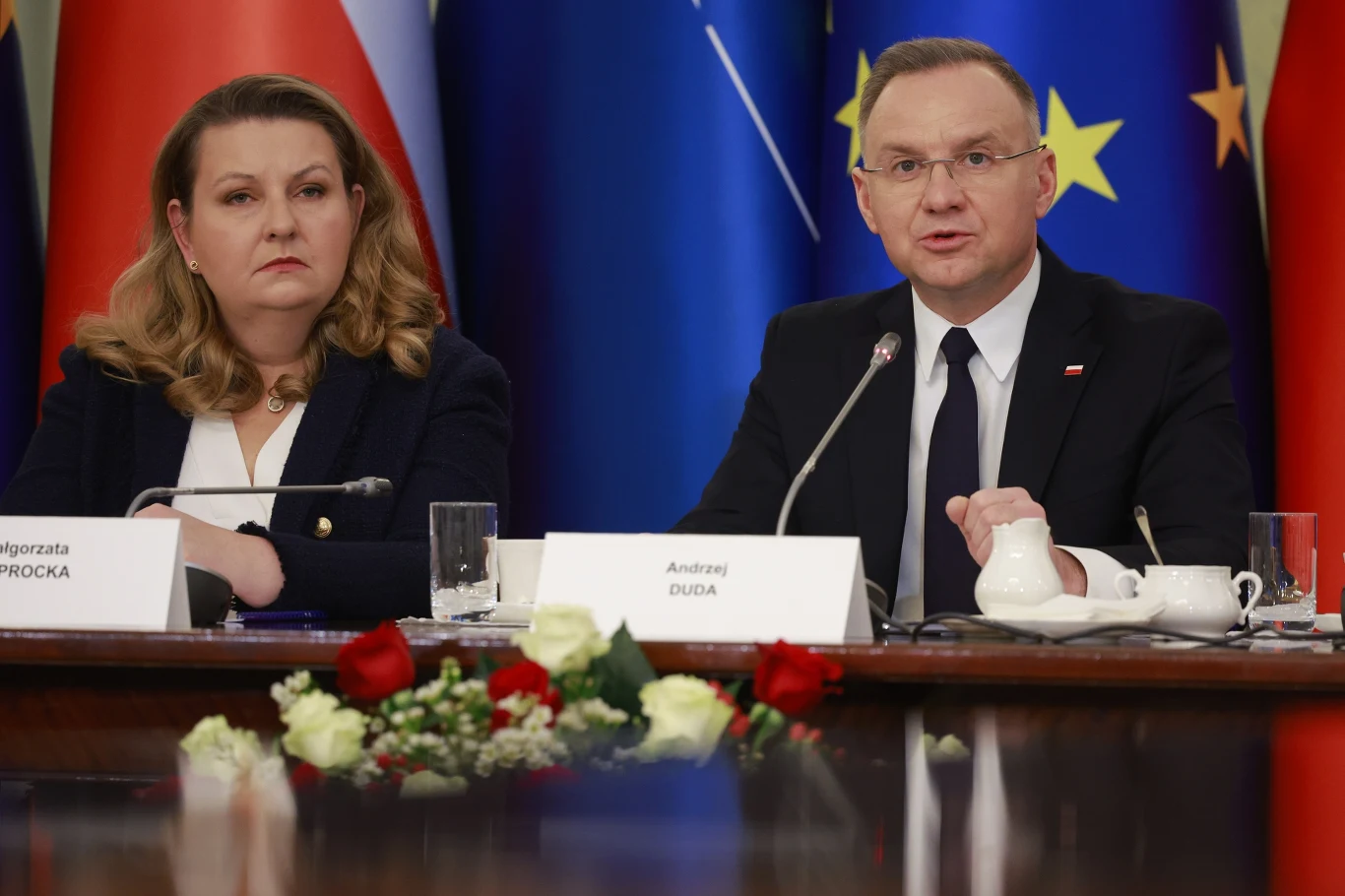 Szefowa kancelarii Andrzeja Dudy zapowiada odejście Dwóch urzędników państwowych siedzi przy stole konferencyjnym, za którym stoją flagi Polski i Unii Europejskiej. W centralnej części stołu leżą biało-czerwone kwiaty, przed osobami znajdują się mikrofony i tabliczki z ich nazwiskami.