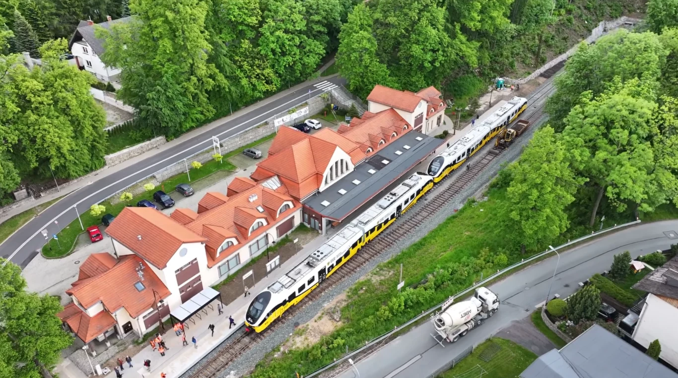 Pociągi wróciły do Karpacza. To nie był udany start. Stacja kolejowa otoczona zielenią, peron z grupą ludzi, nowoczesny pociąg stojący na torach oraz charakterystyczny budynek dworca z czerwonym dachem, widok z lotu ptaka.