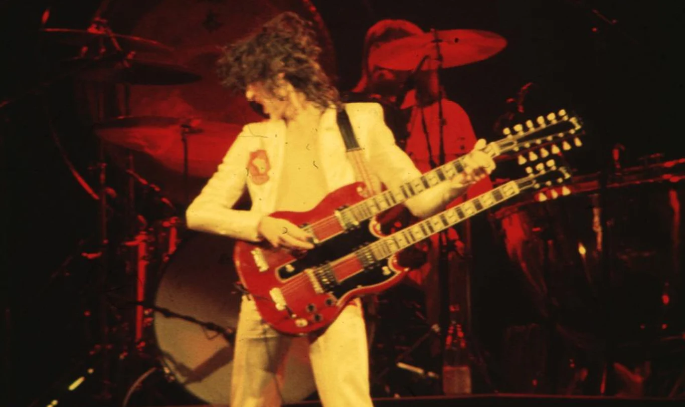 Jimmy Page podczas koncertu Led Zeppelin Muzyk w jasnym garniturze gra na czerwonej gitarze dwugryfowej na scenie, w tle widoczna perkusja i elementy oświetlenia koncertowego, dynamiczna atmosfera występu muzycznego.