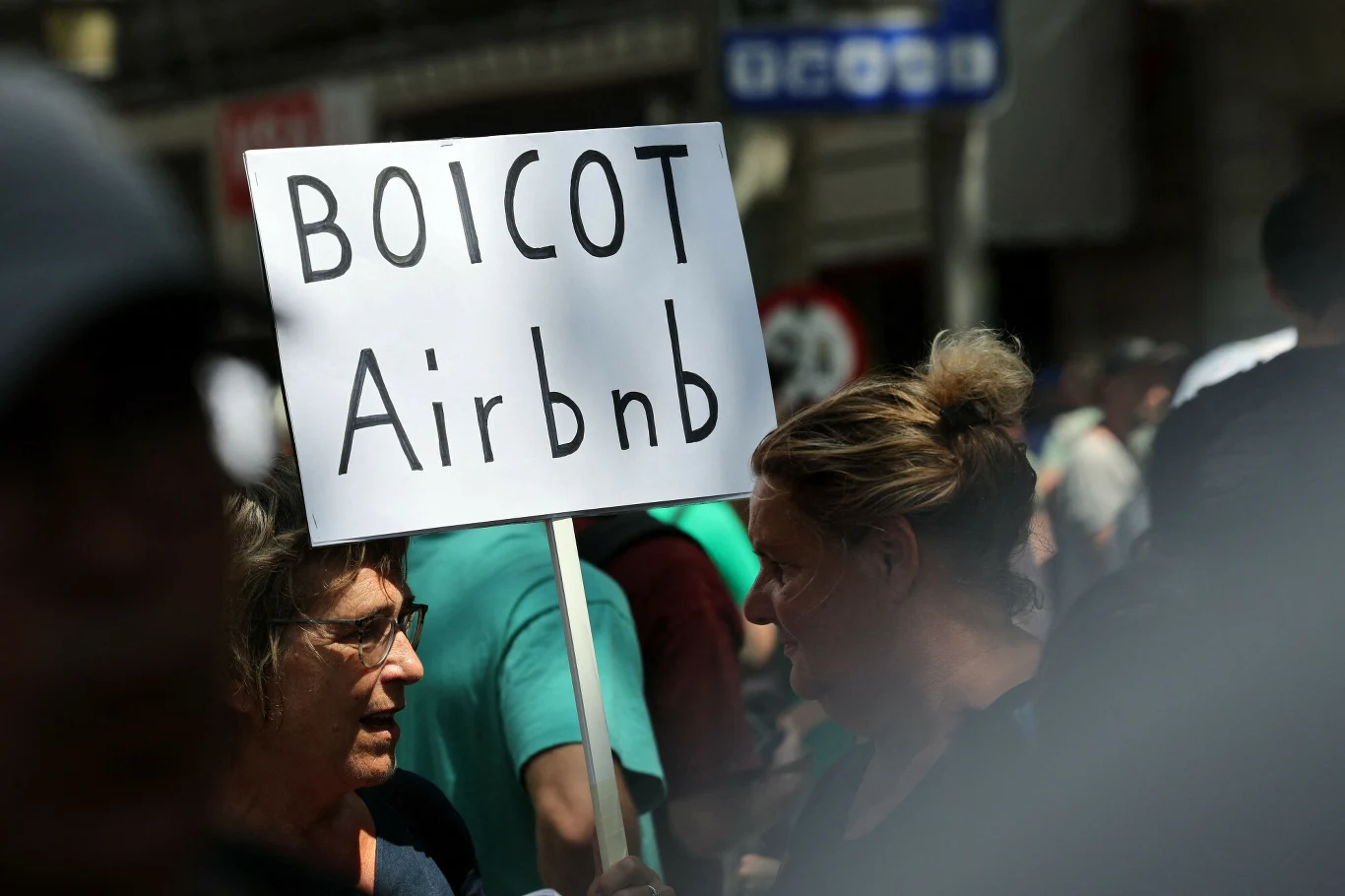 Hiszpanie narzekają na wzmożoną turystykę i protestują Dwie kobiety w tłumie podczas protestu trzymają tablicę z napisem „BOICOT Airbnb”; za nimi widoczne osoby oraz fragmenty budynków miejskich.
