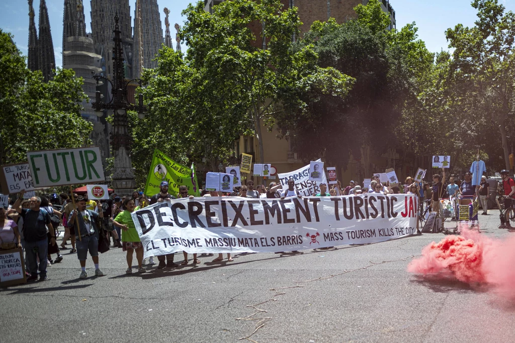 Demonstranci uważają, że turystyka zatruwa im życie Grupa demonstrantów maszeruje ulicą trzymając transparenty i plakaty z hasłami krytykującymi masową turystykę, w tle widoczna charakterystyczna architektura, po prawej stronie unosi się różowy dym.