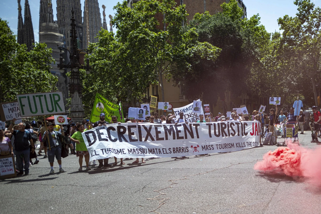 Demonstranci uważają, że turystyka zatruwa im życie Grupa demonstrantów maszeruje ulicą trzymając transparenty i plakaty z hasłami krytykującymi masową turystykę, w tle widoczna charakterystyczna architektura, po prawej stronie unosi się różowy dym.