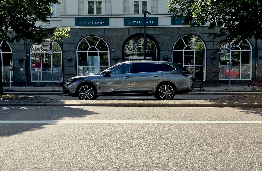 Volkswagen aktualnie oferuje 5 hybryd phev Srebrny samochód osobowy zaparkowany przy ulicy na tle budynku banku Jyske Bank z charakterystycznymi dużymi łukowatymi oknami, drzewa po obu stronach kadru dają częściowy cień.