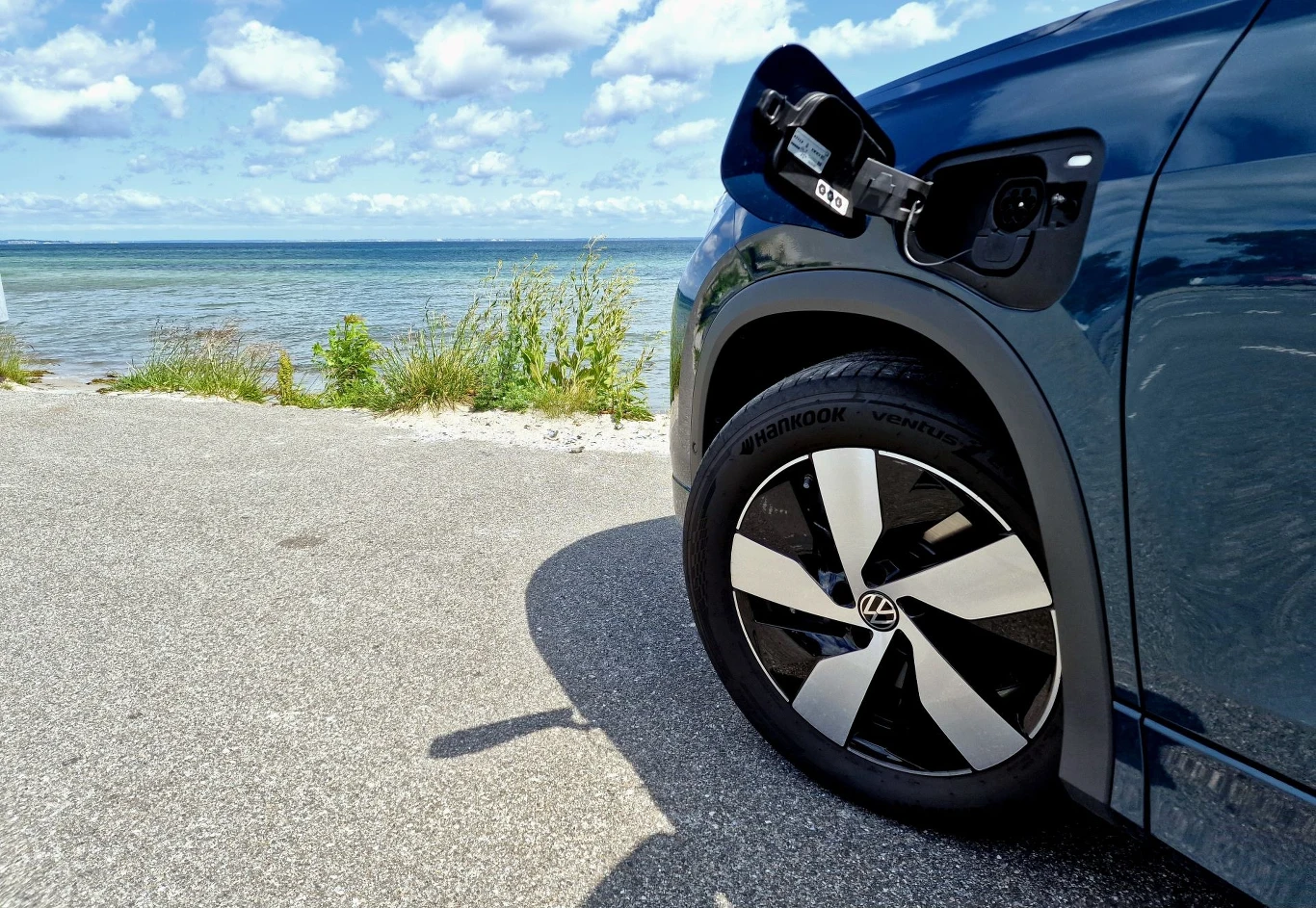 Część elektrycznego samochodu na tle morza i nieba z otwartą klapką od portu ładowania, elegancka felga z logo Volkswagen, asfaltowa droga i roślinność przy linii brzegowej