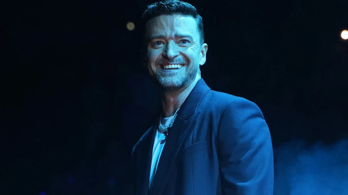 Justin Timberlake wystąpi w Warszawie! Mężczyzna w eleganckim ciemnym garniturze uśmiecha się szeroko, stojąc na tle ciemnego, rozświetlonego punktowymi światłami tła.