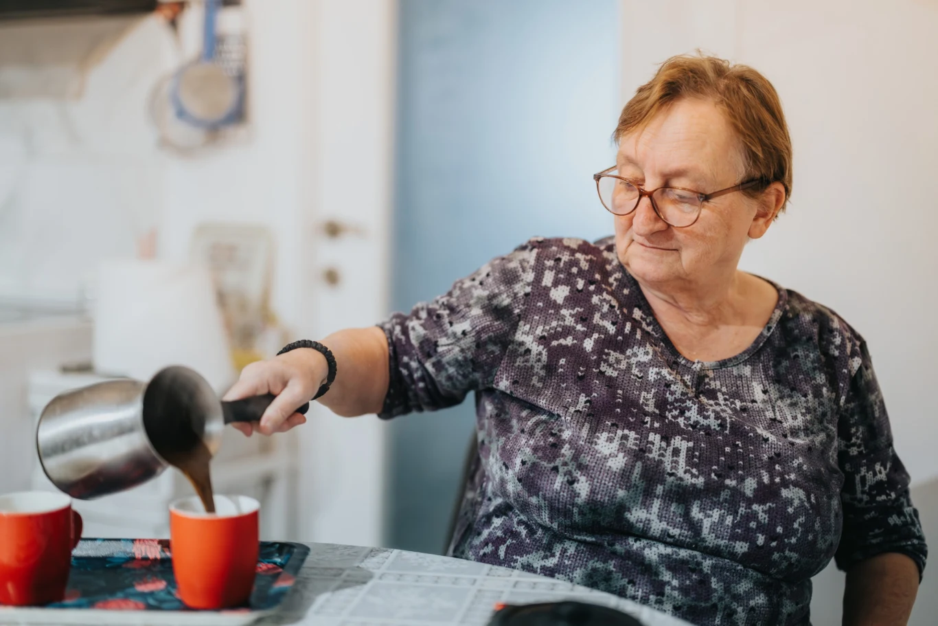Starsza kobieta w okularach nalewa kawę lub herbatę z metalowego dzbanka do czerwonego kubka na stole przykrytym gazetą, obok drugi czerwony kubek.
