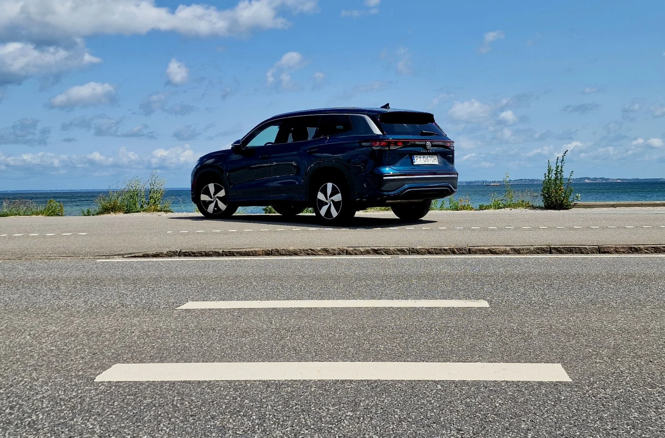 Ciemnoniebieski nowoczesny SUV zaparkowany przy drodze biegnącej wzdłuż wybrzeża, w tle widoczne spokojne morze oraz częściowo zachmurzone niebo.