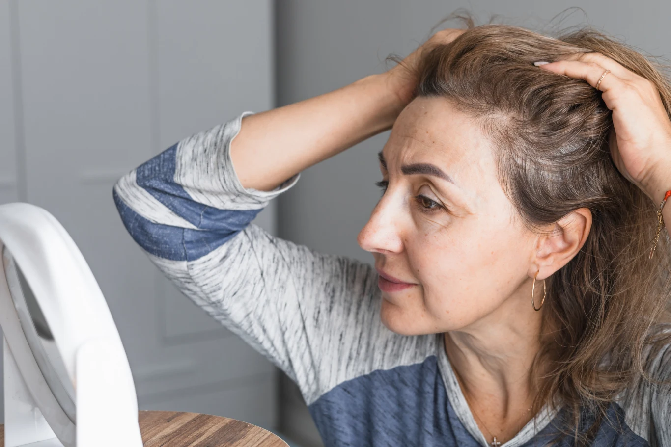 Kobieta w średnim wieku przygląda się swoim włosom w lusterku, trzymając dłońmi włosy na czubku głowy i odsłaniając skronie, przez co eksponuje linię włosów i skórę głowy.