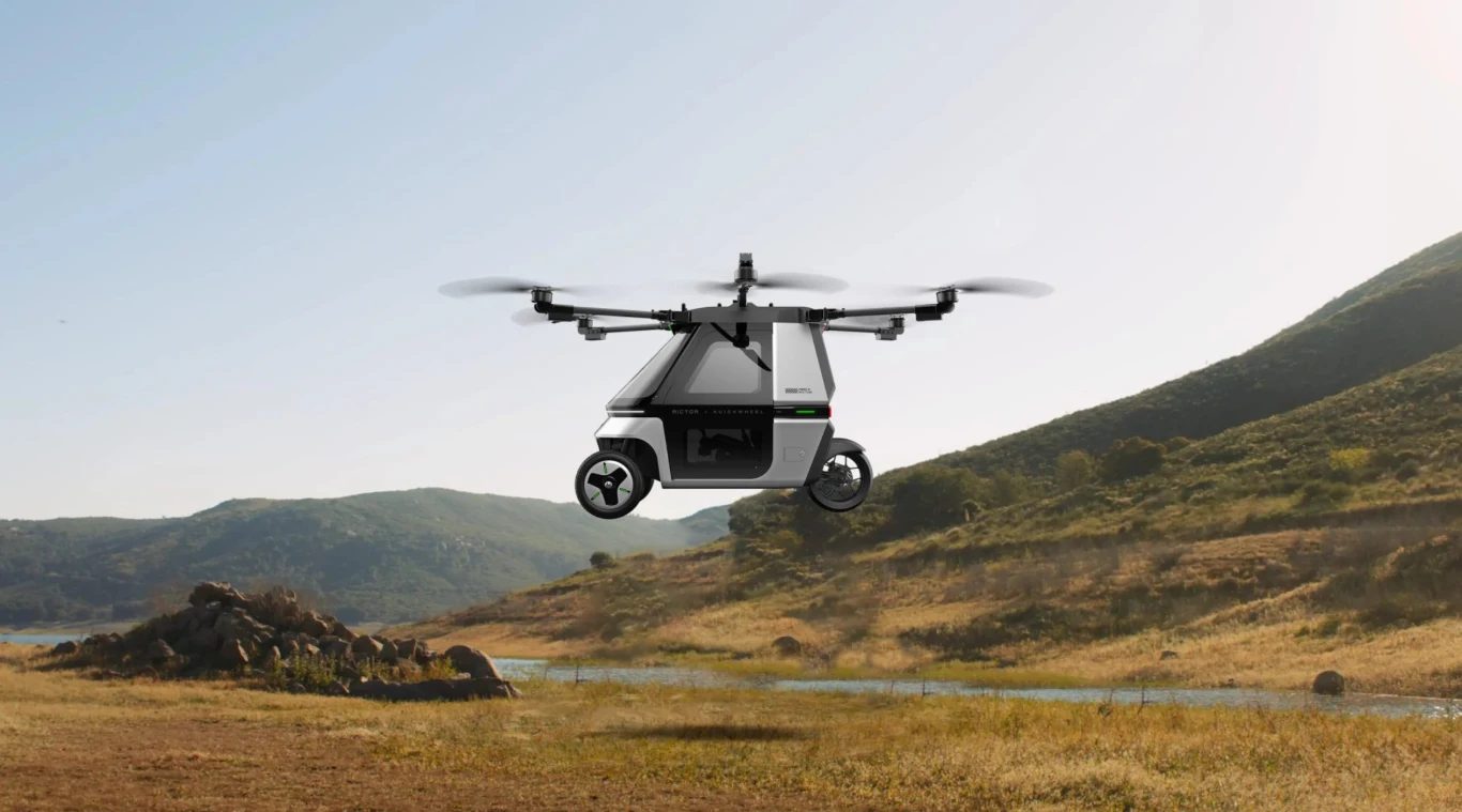 Skyrider X6 w powietrzu. Hybryda motocykla i drona dostępna w przedsprzedaży Nowoczesny dron przypominający mały pojazd unoszący się nisko nad suchym, trawiastym terenem z pagórkowatym krajobrazem oraz niewielką ilością zieleni i wodą w tle.