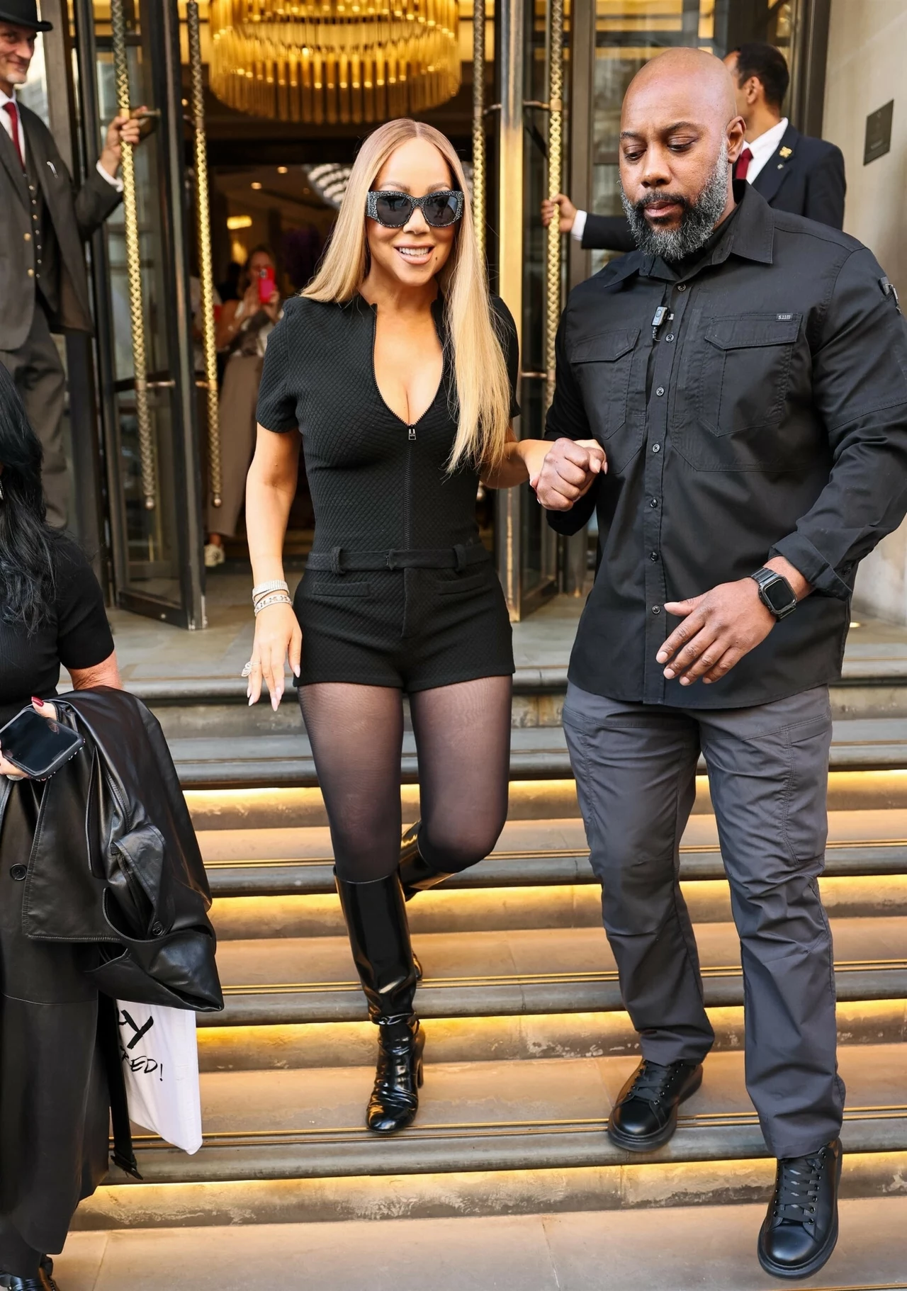 Mariah Carey w wieku 56-lat przeżywa drugą młodość Kobieta w czarnych okularach przeciwsłonecznych i modnym stroju wychodzi z luksusowego budynku po schodach, trzymając się za rękę mężczyzny w ciemnym ubraniu, w tle widać bogato zdobione drzwi obrotowe i eleganckie wnętrze.