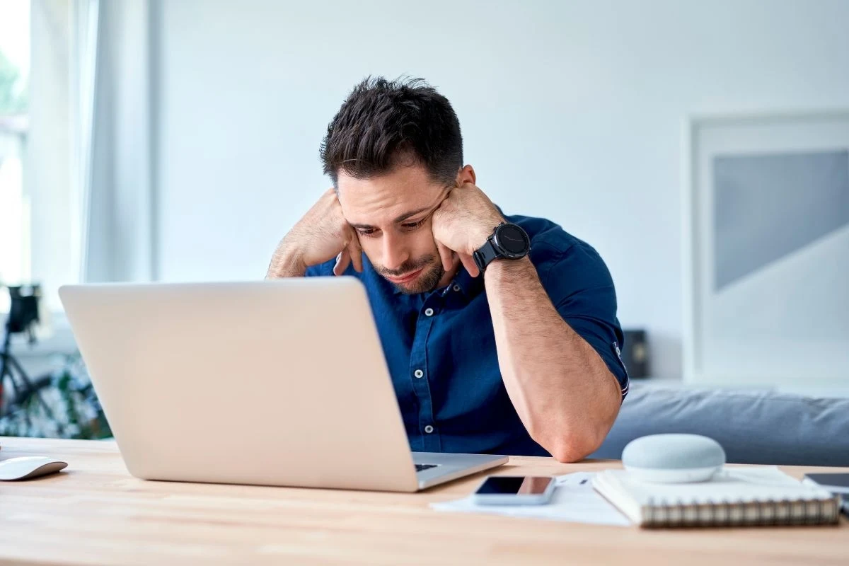 Ekspert uważa, że odrzucony przez kobiety mężczyzna ucieka w internetowe treści mizoginistyczne Mężczyzna w niebieskiej koszuli siedzi przy biurku, wspiera głowę na rękach i patrzy zmartwiony na ekran laptopa. Na biurku obok znajduje się głośnik bezprzewodowy, telefon i notes.