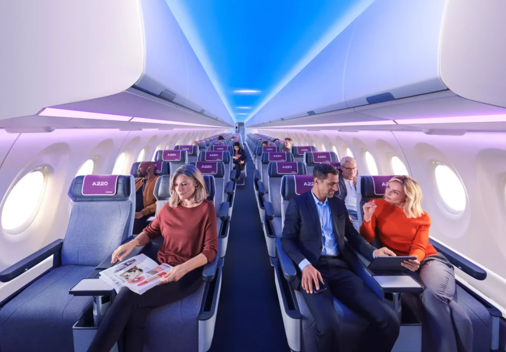 Kabina Airbusa A220. Wnętrze samolotu pasażerskiego klasy ekonomicznej, rzędy wygodnych siedzeń, kilku pasażerów odpoczywających, rozmawiających lub czytających, jasne nowoczesne oświetlenie i spokojna atmosfera lotu.