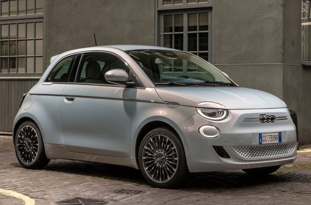 Nowy Fiat 500e, dostosowany do współpracy z modułowymi bateriami firmy Ample, działa w oparciu o automatyczny system wymiany baterii. Nowoczesny, jasnoszary samochód Fiat 500 zaparkowany przy brukowanej ulicy na tle szarego budynku z dużymi oknami, wyróżniający się opływową sylwetką i charakterystycznymi, designerskimi felgami.