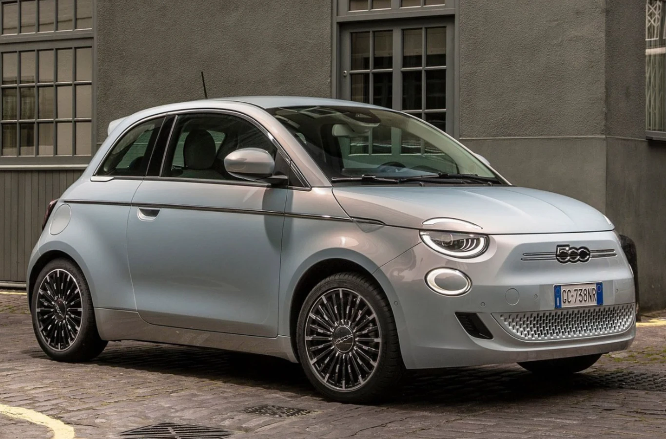 Nowoczesny, jasnoszary samochód Fiat 500 zaparkowany przy brukowanej ulicy na tle szarego budynku z dużymi oknami, wyróżniający się opływową sylwetką i charakterystycznymi, designerskimi felgami.