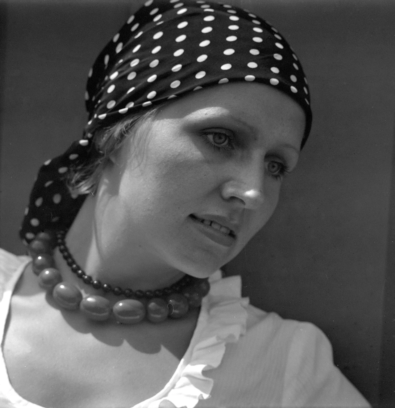 Ewa Śnieżanka podczas Festiwalu w Opolu w 1972 r. Kobieta z delikatnym makijażem, ubrana w białą bluzkę z falbaną i charakterystyczną chustkę w białe grochy na głowie, na szyi dwa naszyjniki – duże korale i mniejsze paciorki, spojrzenie skierowane w bok, wyraz twarzy refleksyjny.
