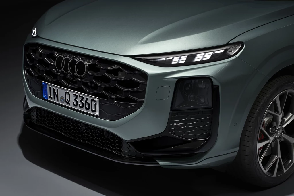 Z przodu nowe Audi Q3 zwraca na siebie uwagę większym grillem i ostrzej narysowanymi, węższymi światłami. Przednia część samochodu marki Audi z charakterystycznym grillem, nowoczesnymi reflektorami LED oraz widoczną niemiecką tablicą rejestracyjną, fragment aluminiowej felgi i maski samochodu w zielonym metalicznym kolorze.