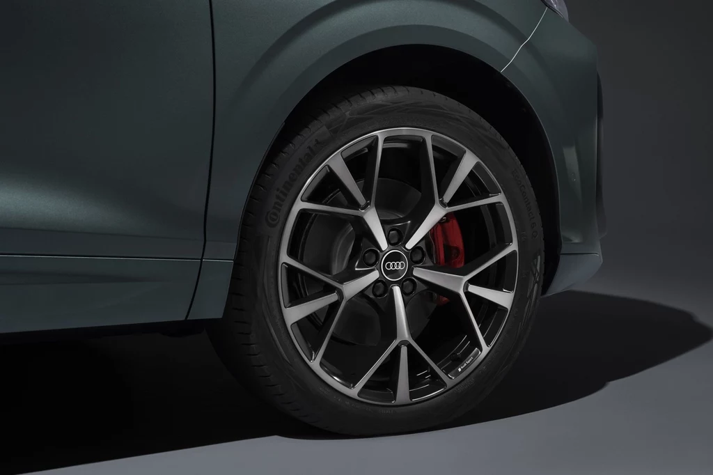 Audi Q3 może stać na felgach w rozmiarach od 17 do 20 cali. Koło samochodowe z charakterystyczną felgą aluminiową o sportowym wzorze, czerwonym zaciskiem hamulcowym i widocznym logo Audi, fragment karoserii pojazdu w kolorze ciemnozielonym.
