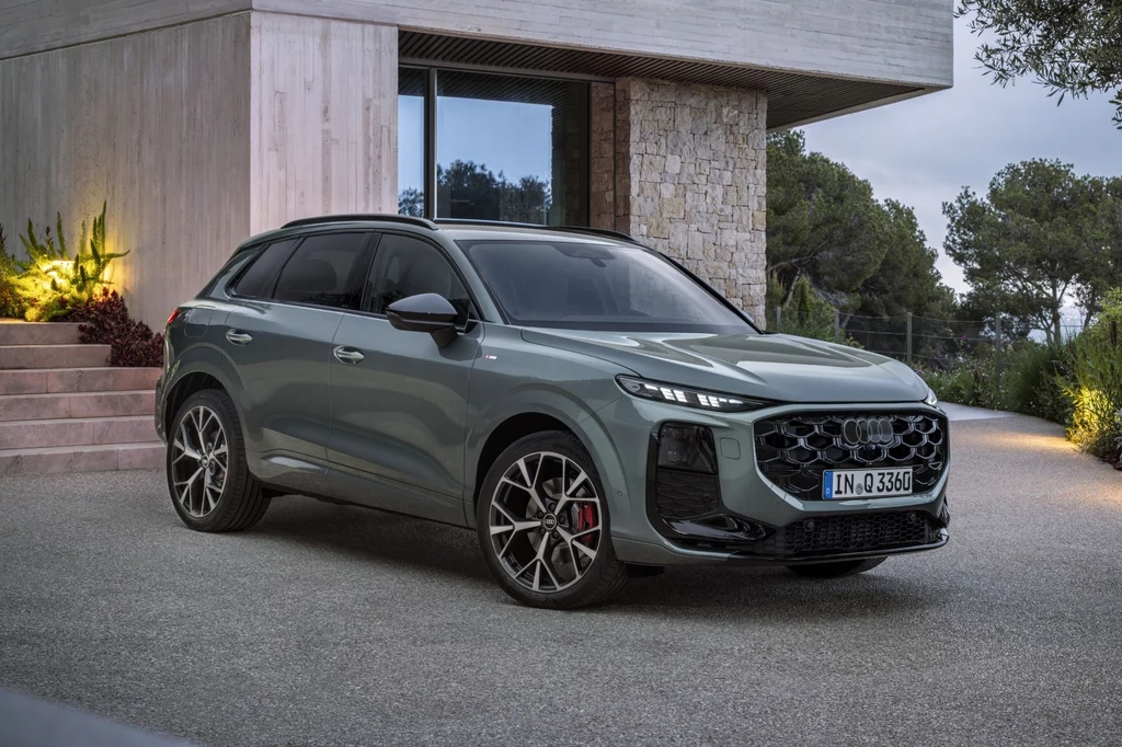 W ofercie Audi Q3, oprócz napędów bazujących na silnikach benzynowych, w ofercie znajdzie się również wariant wysokoprężny generujący 150 KM i 360 Nm. Nowoczesny SUV w szarym kolorze z wyrazistymi, sportowymi felgami, zaparkowany przed minimalistycznym budynkiem z kamienia i betonu, otoczonym zielenią i delikatnie oświetlonym w tle.