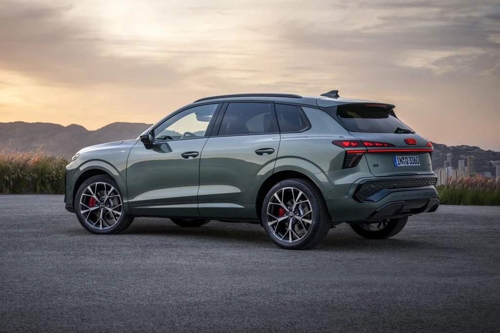 Ceny Audi Q3 w Niemczech zaczynają się od 44 600 euro (ok. 190 tys. zł). Luksusowy SUV w kolorze szarym zaparkowany na pustym placu, widoczny z tyłu na tle gór oraz zachodzącego słońca; auto wyróżnia się sportowymi felgami i czerwonymi zaciskami hamulców.