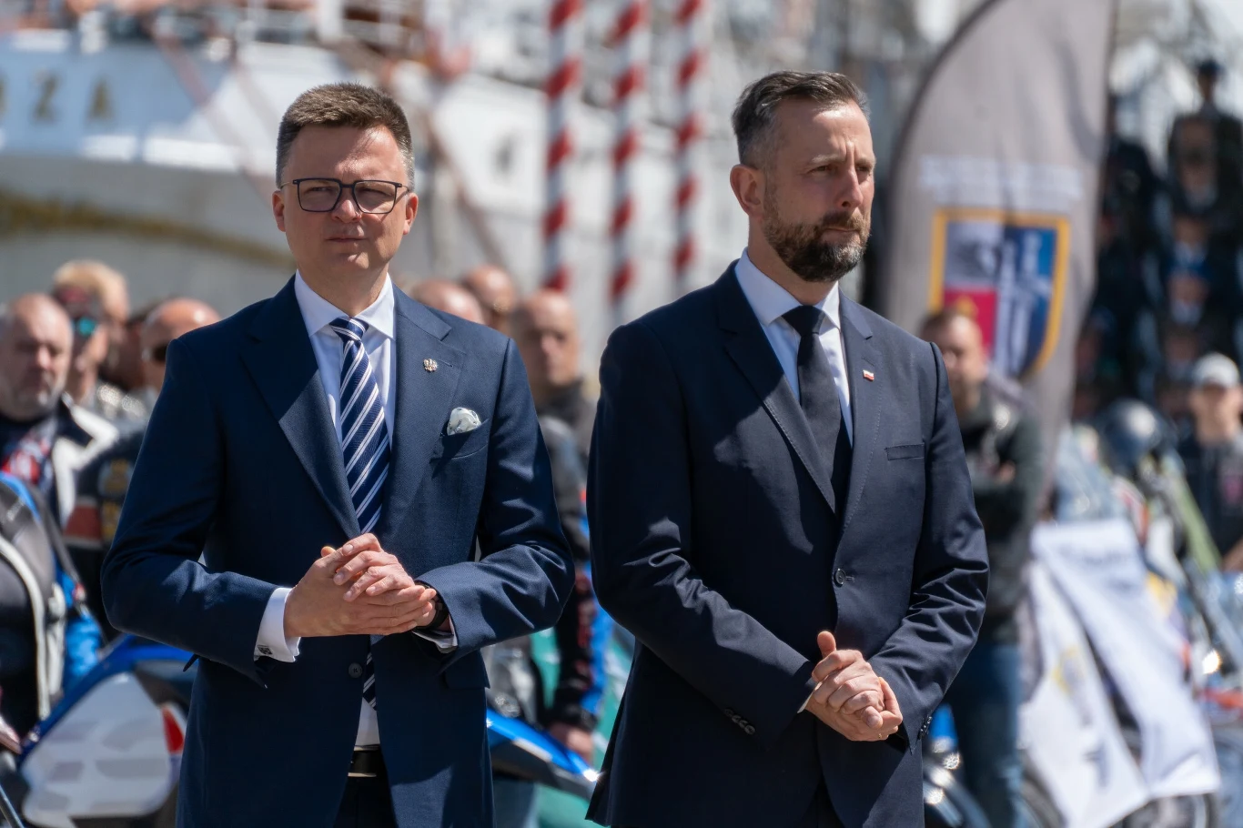 Liderzy Trzeciej Drogi komentują wyniki najnowszego sondażu Dwaj mężczyźni w garniturach stoją obok siebie na tle grupy ludzi i fragmentów biało-czerwonych flag, w tle widoczny fragment masztów i baner reklamowy.