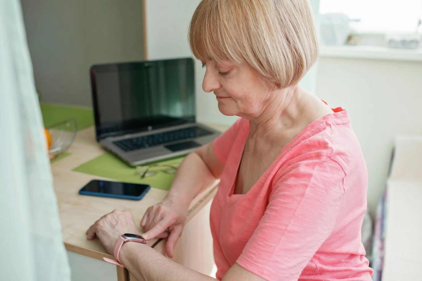 Starsza kobieta w różowej bluzce siedzi przy stole, sprawdzając czas na smartwatchu, w tle znajduje się otwarty laptop oraz smartfon leżący na blacie.