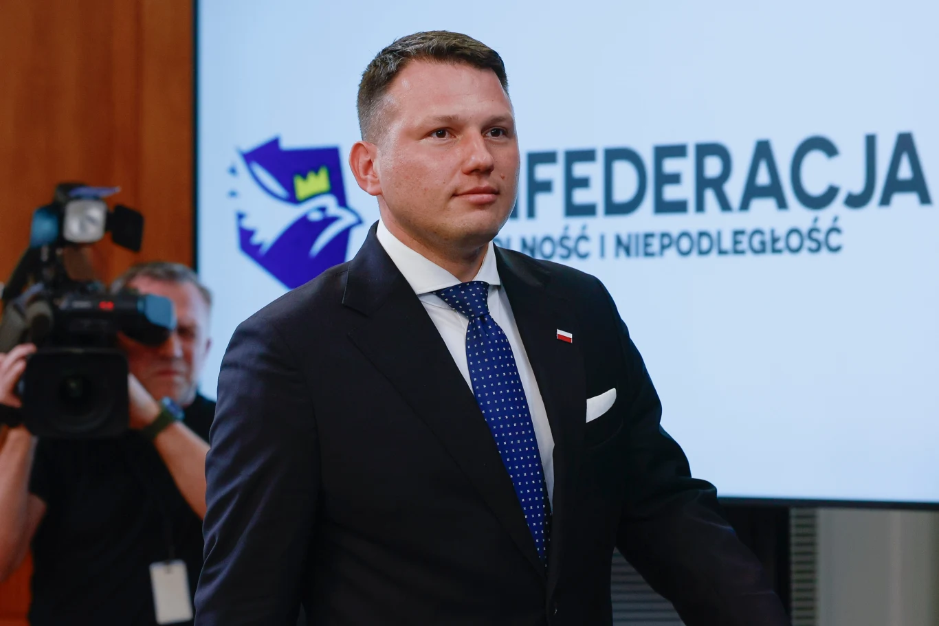Prezes partii Nowa Nadzieja Sławomir Mentzen Mężczyzna w garniturze z przypiętą flagą Polski, stojący przed tablicą z napisem FEDERACJA Stylność i Niepodległość, w tle kamerzysta rejestrujący wydarzenie.