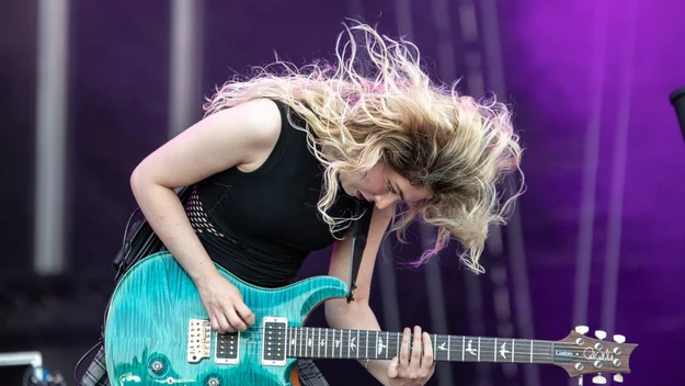 Młoda kobieta z długimi, falującymi włosami energicznie gra na niebieskiej gitarze elektrycznej na scenie, ubrana w czarną bluzkę i spódnicę w kratę, dynamiczne oświetlenie i ruch podkreślają koncertową atmosferę.