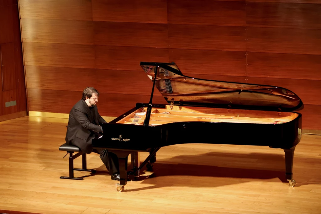 Charles Richard-Hamelin Mężczyzna w garniturze gra na czarnym fortepianie koncertowym na drewnianej scenie przed drewnianą ścianą.