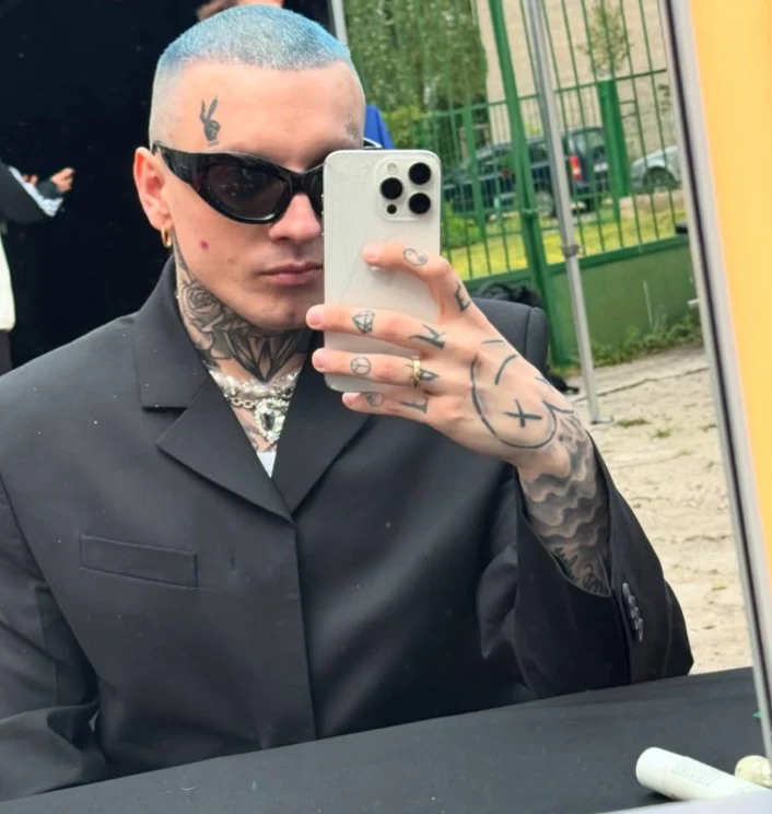 Mężczyzna z niebieskimi włosami wykonuje selfie przy lustrze, ma na sobie ciemny garnitur, nosi duże okulary przeciwsłoneczne, ma widoczne tatuaże na twarzy i dłoniach, w tle widoczne ogrodzenie i fragment budynku.