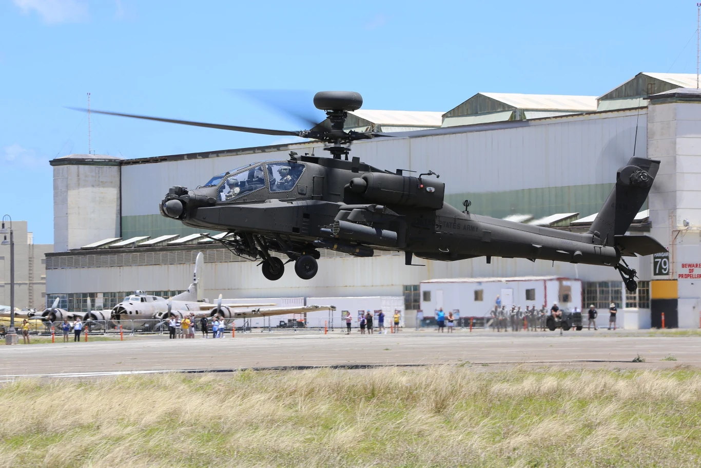 Helikopter bojowy w ciemnym malowaniu unosi się nisko nad pasem startowym przed hangarami lotniczymi, w tle widoczna jest zorganizowana przestrzeń lotniska z kilkoma samolotami i grupami ludzi obserwujących wydarzenie.