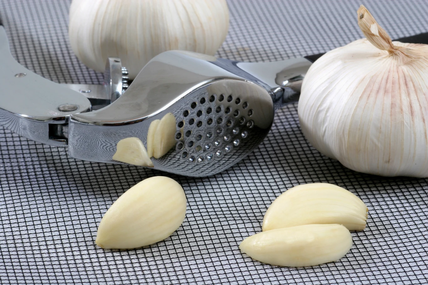 Popularny produkt kuchenny wycofany ze sprzedaży (zdj. ilustracyjne) Metalowa wyciskarka do czosnku obok świeżych ząbków i główek czosnku na kratkowanej powierzchni.