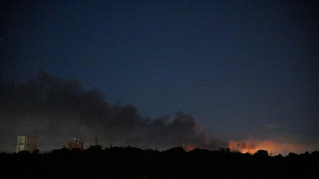 Kłęby dymu nad Kijowem po rosyjskim nocnym ostrzale Nocne niebo nad miastem z widocznymi wieżowcami oświetlonymi światłem oraz łuną i dymem unoszącym się z pożaru lub eksplozji na horyzoncie.