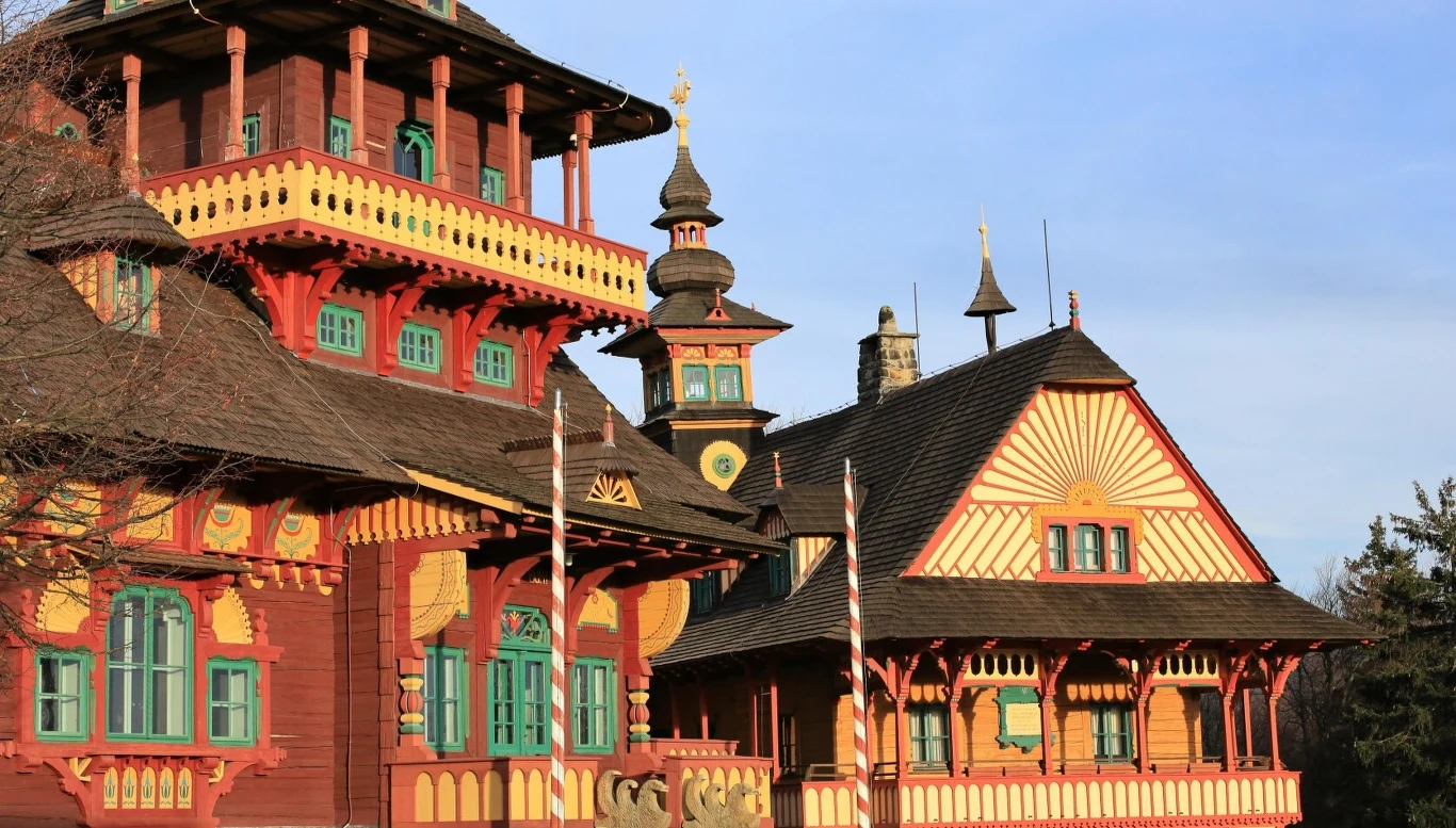 Czechy. Kompleks schronisk na przełęczy Pustevny Kolorowy, drewniany budynek z bogato zdobioną elewacją w stylu zakopiańskim, liczne detaliczne ornamenty, wieżyczki i zielone okna, słoneczna pogoda, niebieskie niebo w tle.