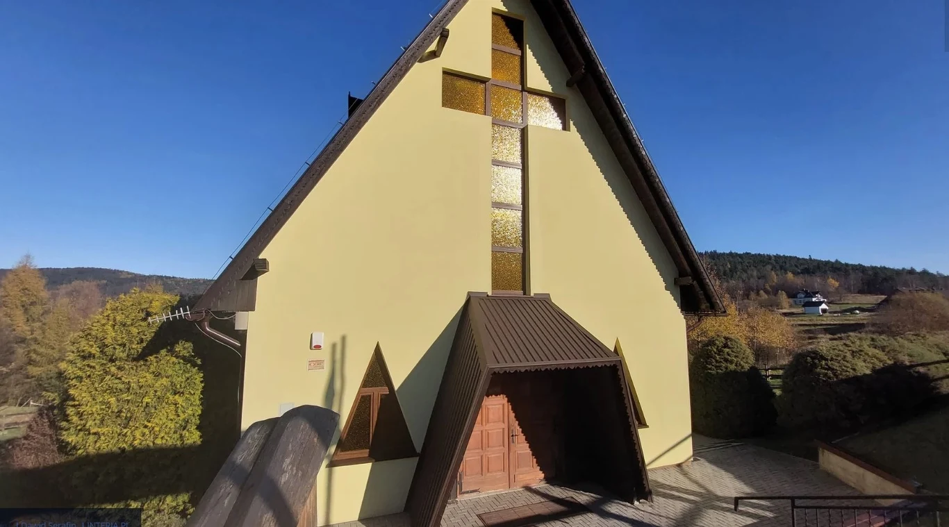 Budynek kościoła z charakterystycznym wysokim dachem i dużym krzyżem z witrażu na frontowej ścianie, otoczony drzewami i położony na tle pagórkowatego krajobrazu z domami w oddali.