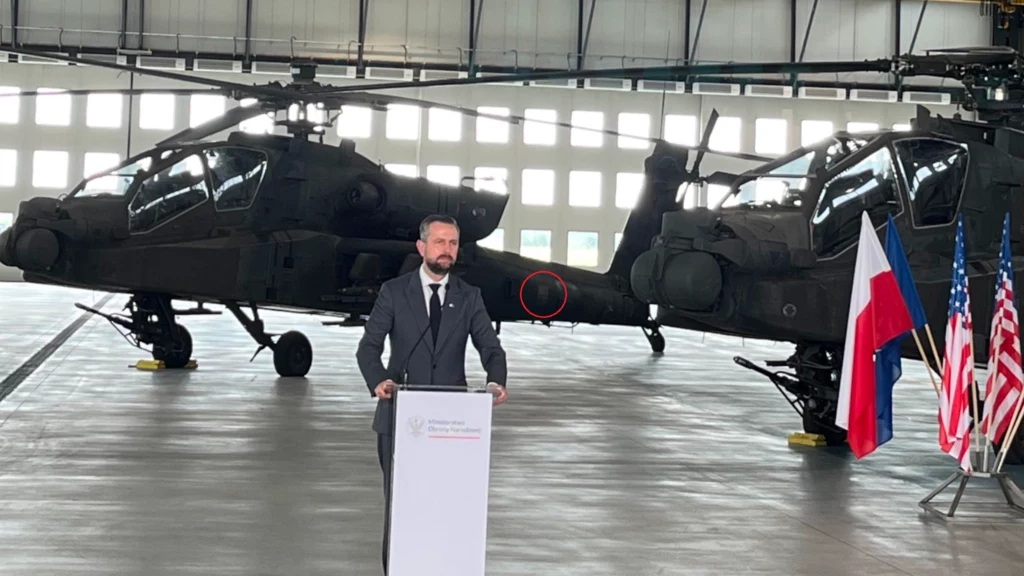 Władysław Kosiniak-Kamysz na uroczystości przekazania pierwszych AH-64D dla żołnierzy 56. Bazy Lotniczej. W tle widać jeden ze śmigłowców z szachownicą low-vis Mężczyzna w garniturze przemawia przy mównicy w hangarze, w tle stoją dwa wojskowe śmigłowce bojowe, obok mównicy znajdują się flagi Polski i Stanów Zjednoczonych.