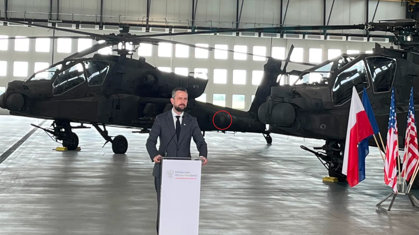 Mężczyzna w garniturze przemawia przy mównicy w hangarze, w tle stoją dwa wojskowe śmigłowce bojowe, obok mównicy znajdują się flagi Polski i Stanów Zjednoczonych.