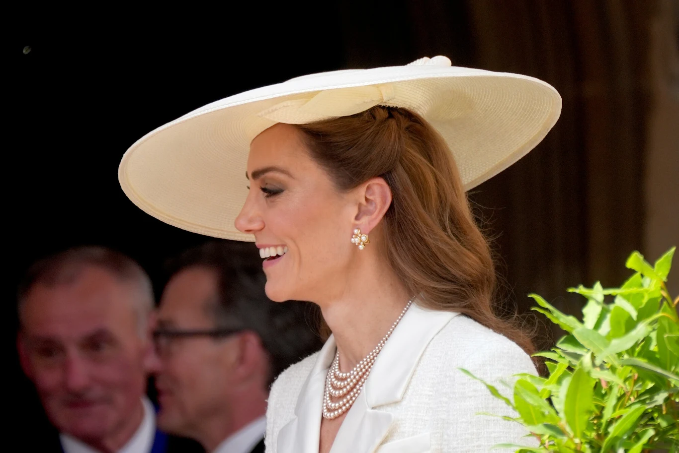 Elegancko ubrana kobieta z długimi, brązowymi włosami, uśmiechnięta, w dużym, kremowym kapeluszu i perłowej biżuterii, stoi w otoczeniu innych osób, na pierwszym planie widoczne zielone liście.