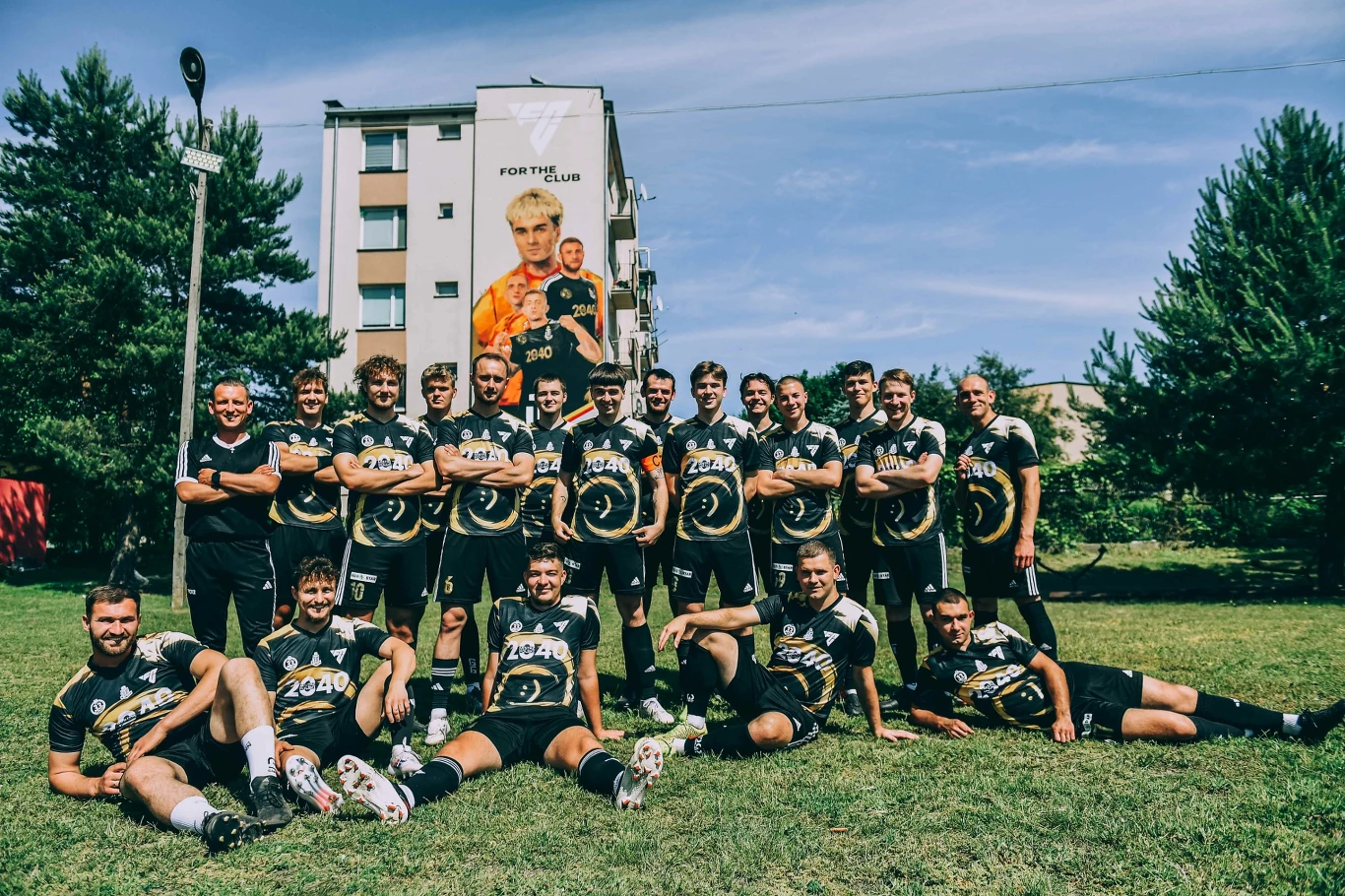 LKS Tajfun Ostrów Lubelski Grupa młodych mężczyzn w sportowych strojach drużyny piłkarskiej pozuje razem na murawie, tworząc zwarty zespół, w tle widoczny blok mieszkalny z dużym banerem reklamowym przedstawiającym sportowców.