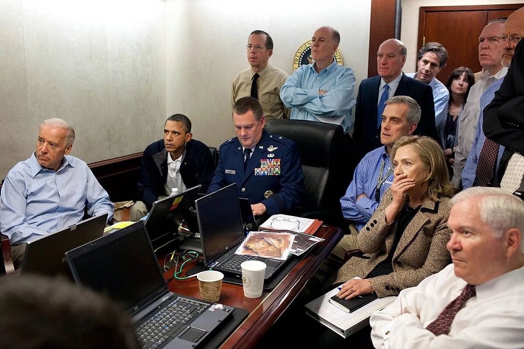 Prezydent USA Barack Obama i wiceprezydent Joe Biden wraz z członkami zespołu ds. bezpieczeństwa narodowego otrzymują aktualne informacje na temat Operacji Neptune Spear , misji przeciwko Osamie bin Ladenowi Grupa wysokich rangą urzędników państwowych oraz wojskowych zgromadzona przy stole konferencyjnym, wszyscy skoncentrowani na ekranach laptopów i monitorów, atmosfera skupienia i napięcia.