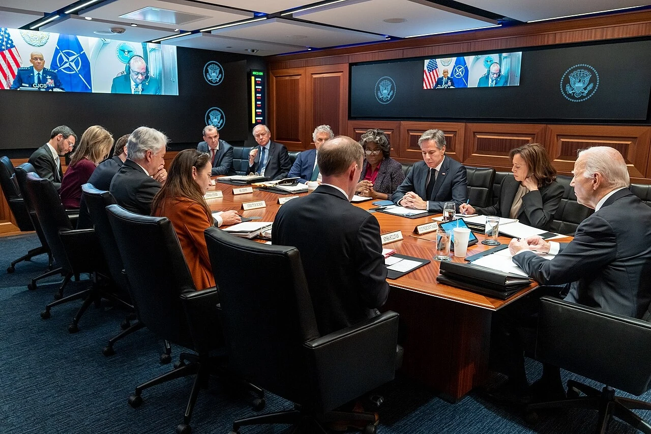 Prezydent Biden i wiceprezydent Harris spotykają się ze swoimi zespołami ds. bezpieczeństwa narodowego w Situation Room Białego Domu 11 października 2023 r. Grupa wysokich rangą urzędników siedzi przy dużym stole konferencyjnym w nowoczesnej sali narad, na ekranach widoczne są symbole NATO i połączenia wideo z innymi uczestnikami spotkania, na stole znajdują się dokumenty, laptopy i napoje.