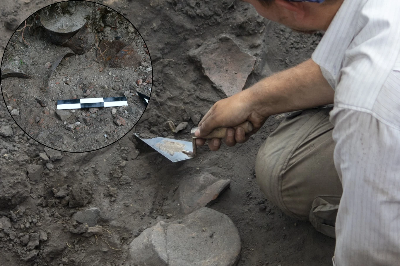 Czasem do odkryć dochodzi ze względu na prowadzenie w danym miejscu inwestycji. We Wrocławiu był to remont nawierzchni. Zdj. ilustracyjne. Ręka archeologa używającego kielni do precyzyjnego oczyszczania fragmentów ceramiki znajdujących się w ziemi, zbliżenie na miejsce wykopaliskowe z fragmentami naczyń oraz linijką pomiarową do skali.