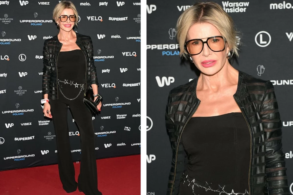 Joanna Racewicz Kobieta w modnym czarnym stroju, czarnych okularach i z czarną kopertówką pozuje na tle ścianki z logotypami podczas wydarzenia branżowego.